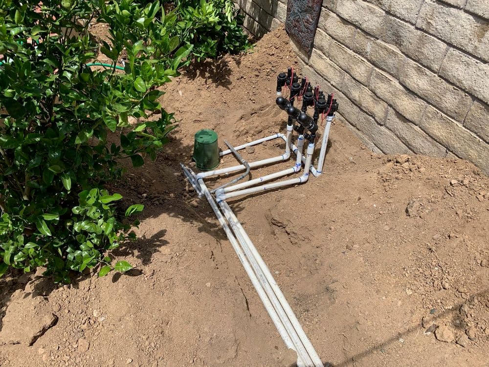 Irrigation system with pipes and valves installed in a garden bed, surrounded by soil and shrubs.