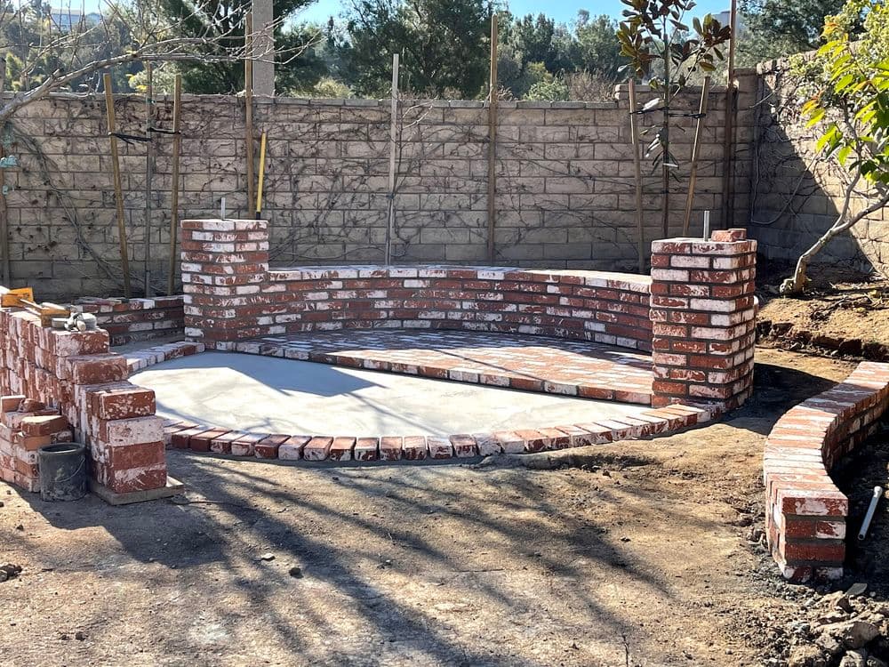 Brick patio under construction with curved seating walls and a natural landscape backdrop.