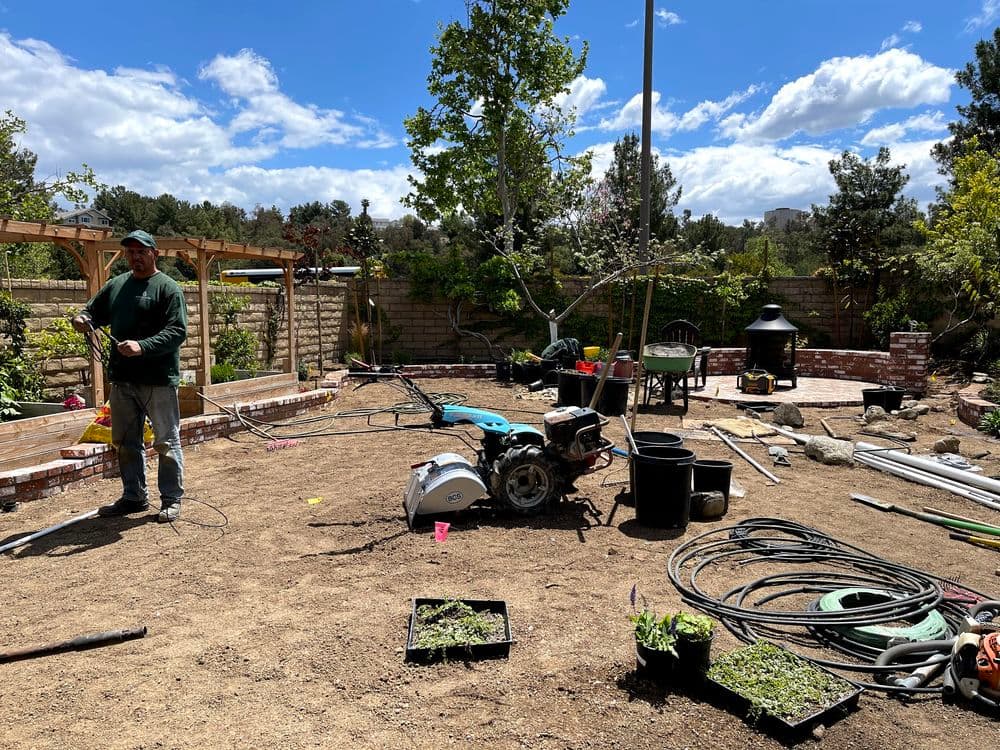 Man working in a garden with tools and plants under a blue sky and clouds.