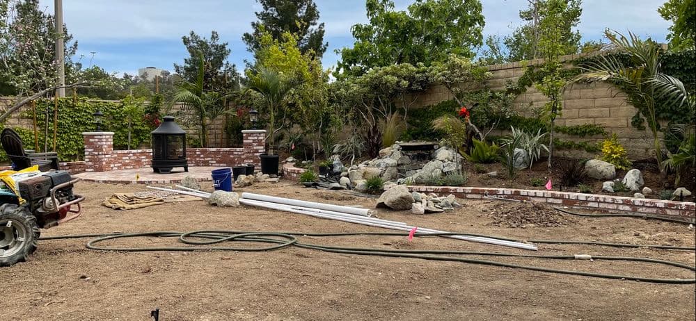 Landscaping project in progress with rocks, plants, and a brick fire pit in a suburban garden.