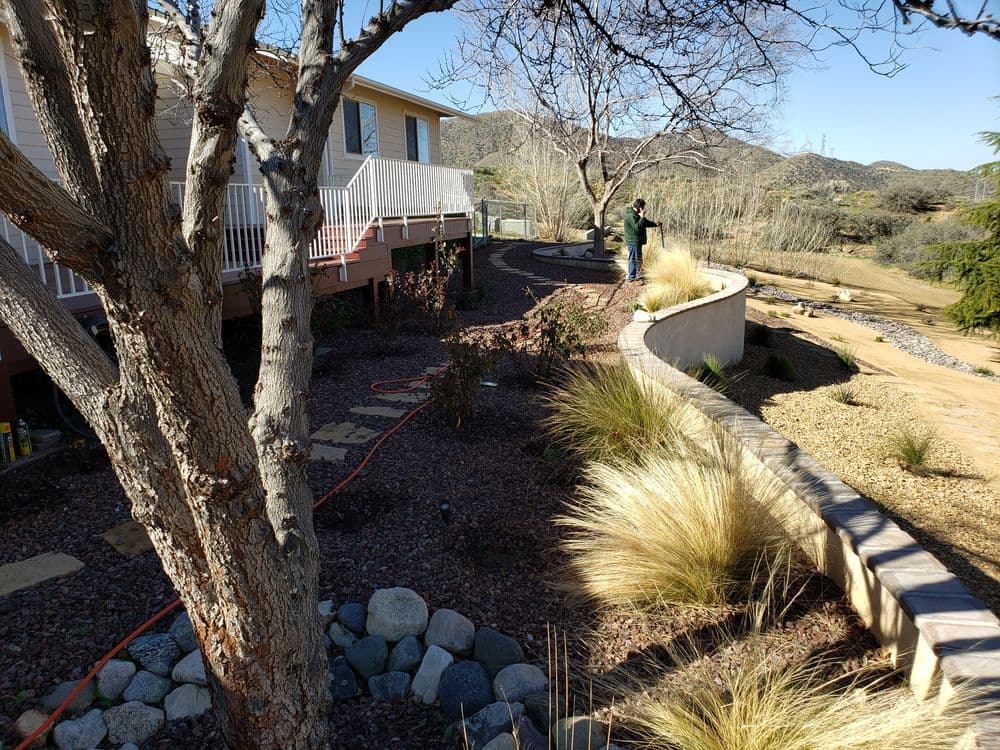 Landscaped yard with stone path, shrubs, and a person gardening near a house in a hilly setting.