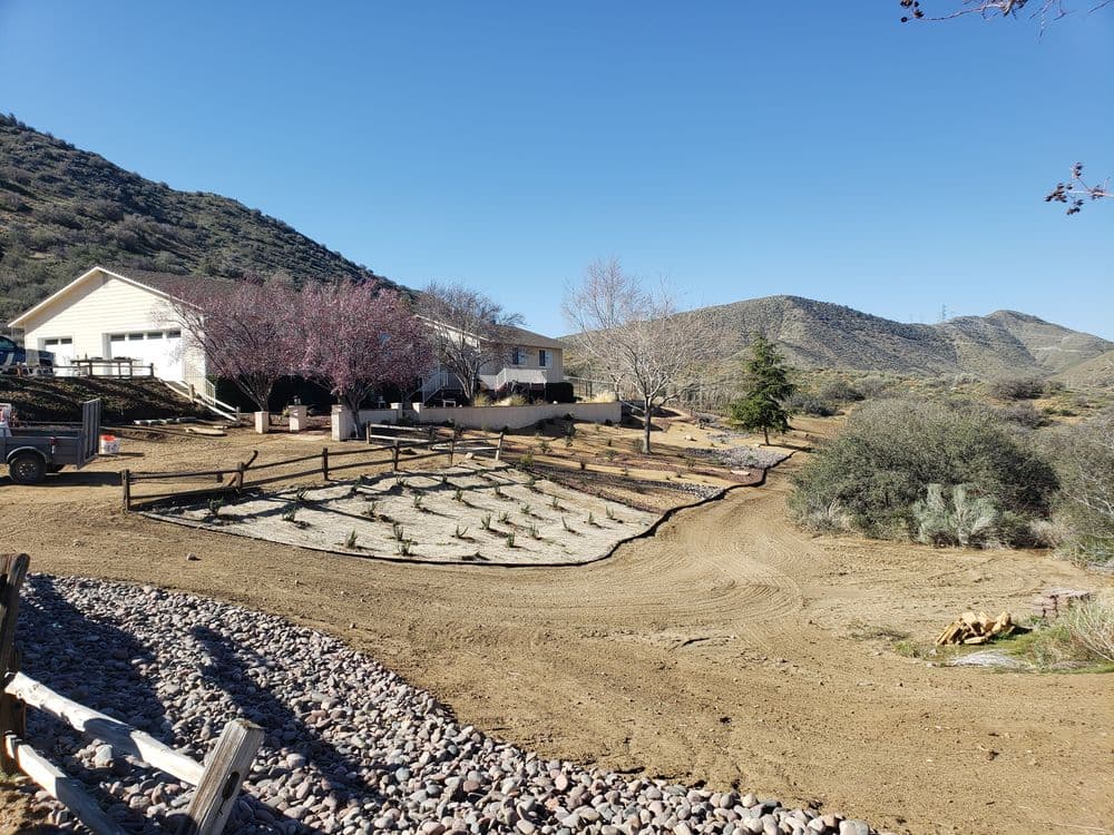 Rural landscape featuring a house, landscaped garden, and mountains under a clear blue sky.
