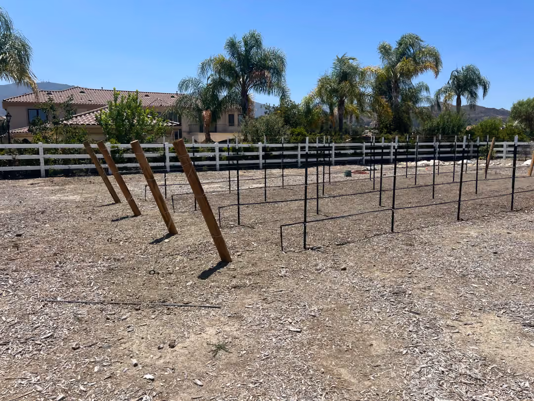 A construction worker builds a wooden fence in a dirt area, with tools and materials visible nearby.